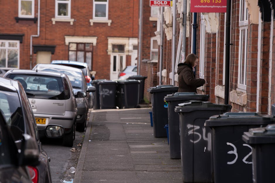 A row of dark grey wheelie bins with closed lids positioned along a narrow urban sidewalk adjacent to a brick residential building. The bins are marked with white spray-painted numbers, and some have
